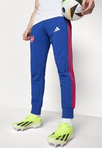 Modré tepláky s červenými pruhy po stranách, s logem Arsenalu. Model má na sobě neonově žluté fotbalové kopačky s černými detaily.