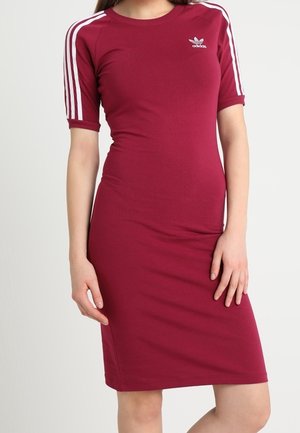 Kobieta ubrana w dopasowaną bordową sukienkę za kolano z białymi paskami Adidas na rękawach i logo na piersi na jednolitym tle.