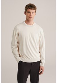 Pull à col rond beige clair en tissu doux, avec poignets et ourlet côtelés, porté sur un pantalon foncé, sur un fond neutre.