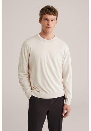 Lichtbeige trui met ronde hals van zachte stof, met geribbelde manchetten en zoom, gedragen over een donkere broek, met een neutrale achtergrond.