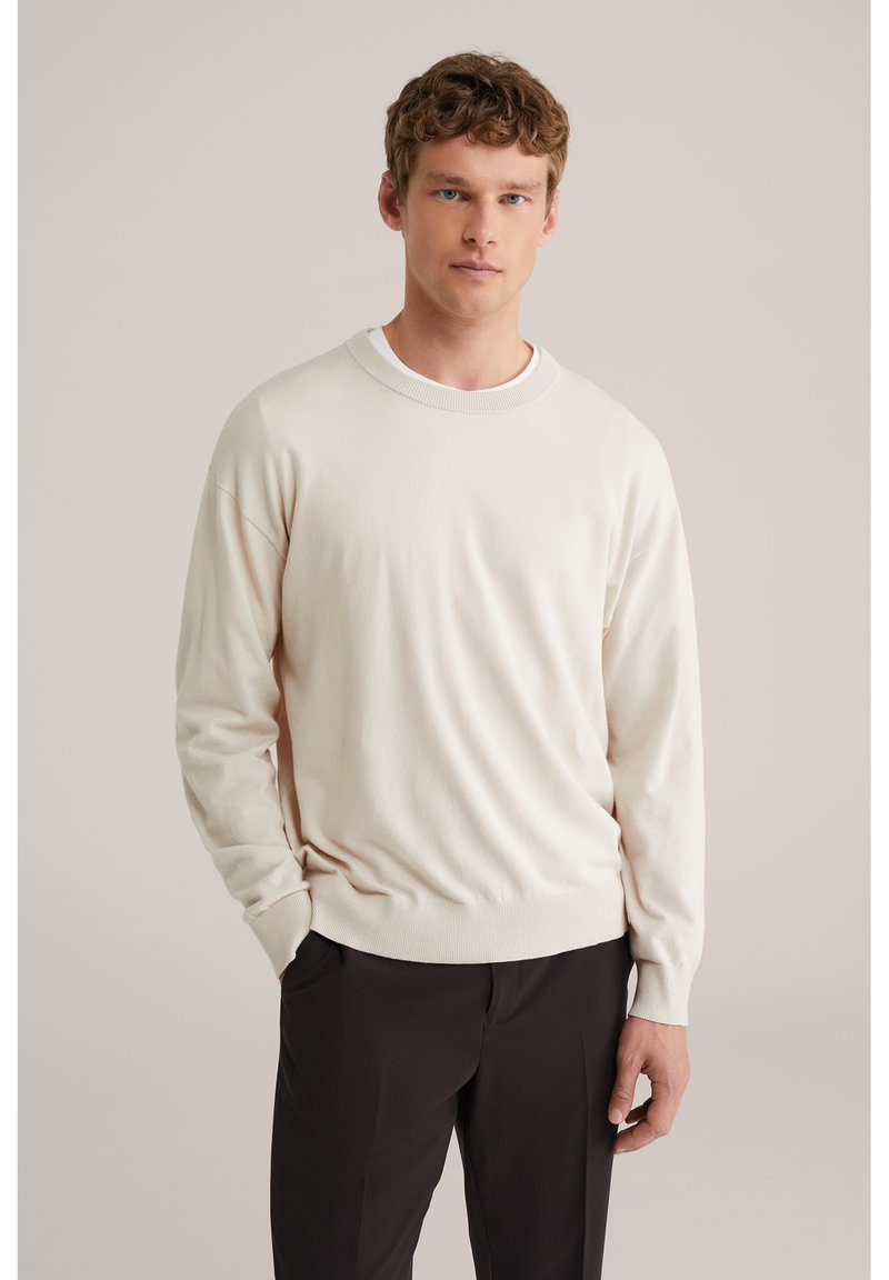 Pull à col rond beige clair en tissu doux, avec poignets et ourlet côtelés, porté sur un pantalon foncé, sur un fond neutre.