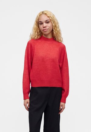 Jeune femme aux cheveux blonds bouclés portant un pull rouge à manches longues et un pantalon noir, debout devant un fond clair uni.