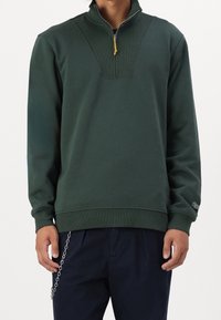 Sweat-shirt vert foncé avec un col zippé demi-lancé, design côtelé au niveau du col et des poignets. Présente un détail de logo sur la manche.
