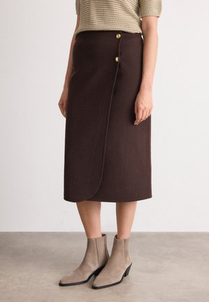 Brun, kneelengde skjørt med omslagdesign og to gullfargede knapper i livet. Båret med beige ankelstøvler på et nøytralt gulv.