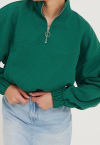 Sudadera de forro polar verde con cremallera de cuarto, que presenta un cuello alto y puños gather. Combinada con una falda de mezclilla azul claro.