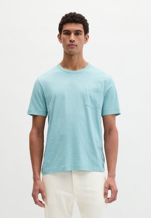 Man draagt een lichtblauw T-shirt met korte mouwen en borstzakje, en witte broek, staand tegen een effen witte achtergrond.