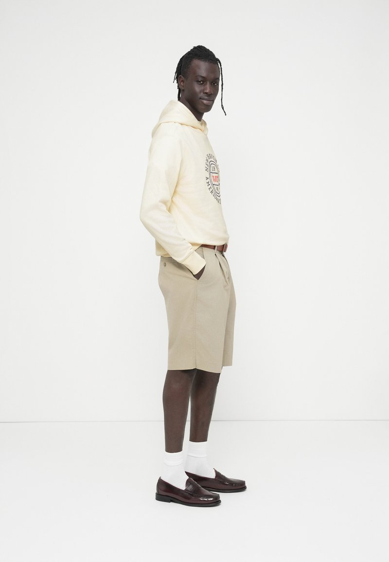 Sweat-shirt beige avec logo, short beige, chaussettes blanches et mocassins bordeaux. Le mannequin est de profil, montrant la tenue et ses détails.