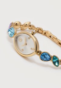 Χρυσό βραχιόλι ρολόι με οβάλ λευκό καντράν, χρυστά δείκτες και διακοσμητικά από πέτρες σε μπλε, τιρκουάζ και μωβ χρώμα πάνω στους κρίκους, σε λευκό φόντο.