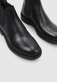 Botas Chelsea de cuero negro con punta afilada, paneles laterales elásticos y suela de goma plana. Textura suave y diseño minimalista.