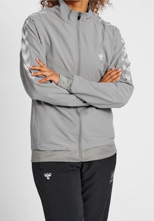 Osoba z założonymi ramionami, ubrana w jasnoszarą kurtkę na zamek błyskawiczny z białymi paskami w kształcie litery V oraz czarne spodnie z logo na udzie.