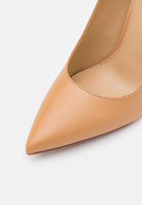 Chaussure à talon haut en cuir beige à bout pointu avec couture visible le long du bord et doublure intérieure lisse sur fond blanc.