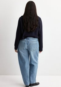 Osoba s dlouhými tmavými vlasy, oblečená v černém tričku s dlouhým rukávem, světle modrých džínách s vysokým pasem a černých botách, stojící zády.