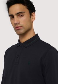 Námořnicky modré polokošile se zeleným akcentem na límci, dvěma knoflíky u rozparku a malým vyšitým logem na prsou. Materiál působí hladce a texturovaně.