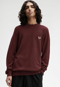 Giovane con capelli ricci che indossa un maglione a maniche lunghe di colore bordeaux con un piccolo logo bianco a forma di alloro sul petto e pantaloni neri.