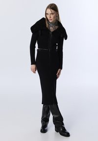Zwarte geribbelde trui jurk met een ritssluiting aan de voorkant, voorzien van een nepkraag van bont. Gecombineerd met glanzende zwarte laarzen tot de knie, waardoor een slanke silhouet ontstaat.