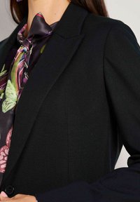 Eine Frau trägt ein schwarzes Blazer über einer blumigen Bluse mit einer gebundenen Schleife am Kragen und passt gerade den Knopf des Blazers an.
