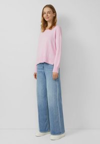 Hellrosa gestrickter Pullover mit einem gerippten Saum, kombiniert mit hellblauen Jeans mit weitem Bein und weißen Turnschuhen, dargestellt vor einem neutralen Hintergrund.