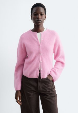 Donna che indossa un cardigan rosa tenue con bottoni sopra una maglietta bianca e pantaloni marrone scuro, in piedi con una mano in tasca contro uno sfondo semplice.