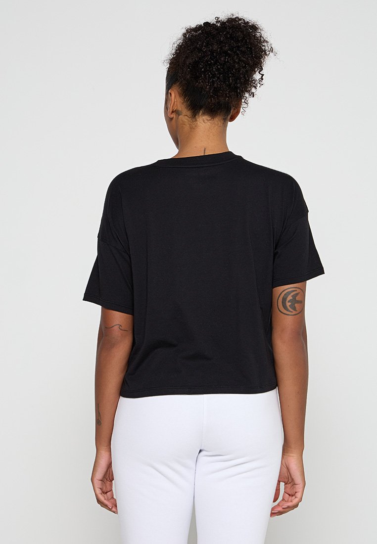 Femme aux cheveux bouclés portant un t-shirt noir à manches courtes et un pantalon blanc, se tenant de dos devant un fond uni.