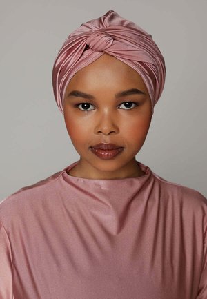 Vrouw met een gladde bruine huid die een glanzende roze tulband en een bijpassende roze satijnen top draagt, kijkt recht in de camera tegen een effen grijze achtergrond.