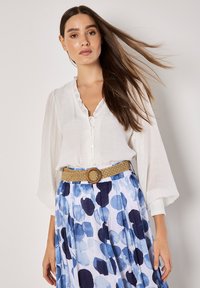 Witte textuurblouse met een gerimpelde halslijn en knoopsluiting, gecombineerd met een blauw waterverfpatroonrok en een geweven riem.