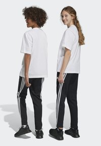 Camisetas de algodón blancas combinadas con pantalones de chándal negros que tienen tres rayas blancas a los lados, y zapatillas deportivas negras. Vista trasera.