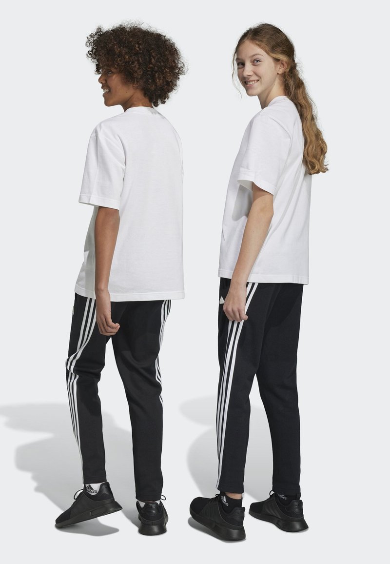 Camisetas de algodón blancas combinadas con pantalones de chándal negros que tienen tres rayas blancas a los lados, y zapatillas deportivas negras. Vista trasera.