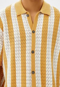 Kortärmad stickad polo med växlande gula och vita zigzag-mönster, knappar och ribbad krage. Lätt textur.