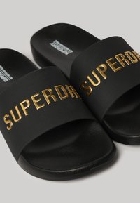 Des sandales noires à glissière avec une sangle texturée ornée du lettrage or "SUPERDRY", une semelle plate et un design minimaliste.