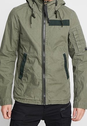 Mann trägt olivgrüne Kapuzenjacke mit schwarzem Reißverschluss und Taschen-Details, kombiniert mit schwarzen Cargohosen, steht mit entspannten Armen.