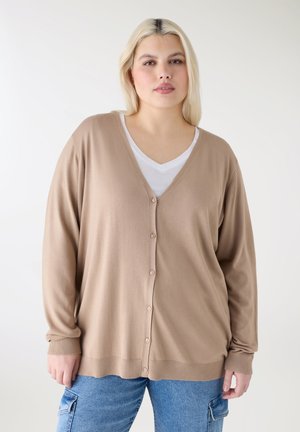 Giovane donna con capelli biondi che indossa un cardigan beige con bottoni sopra una camicia bianca e jeans blu su uno sfondo chiaro e uniforme.