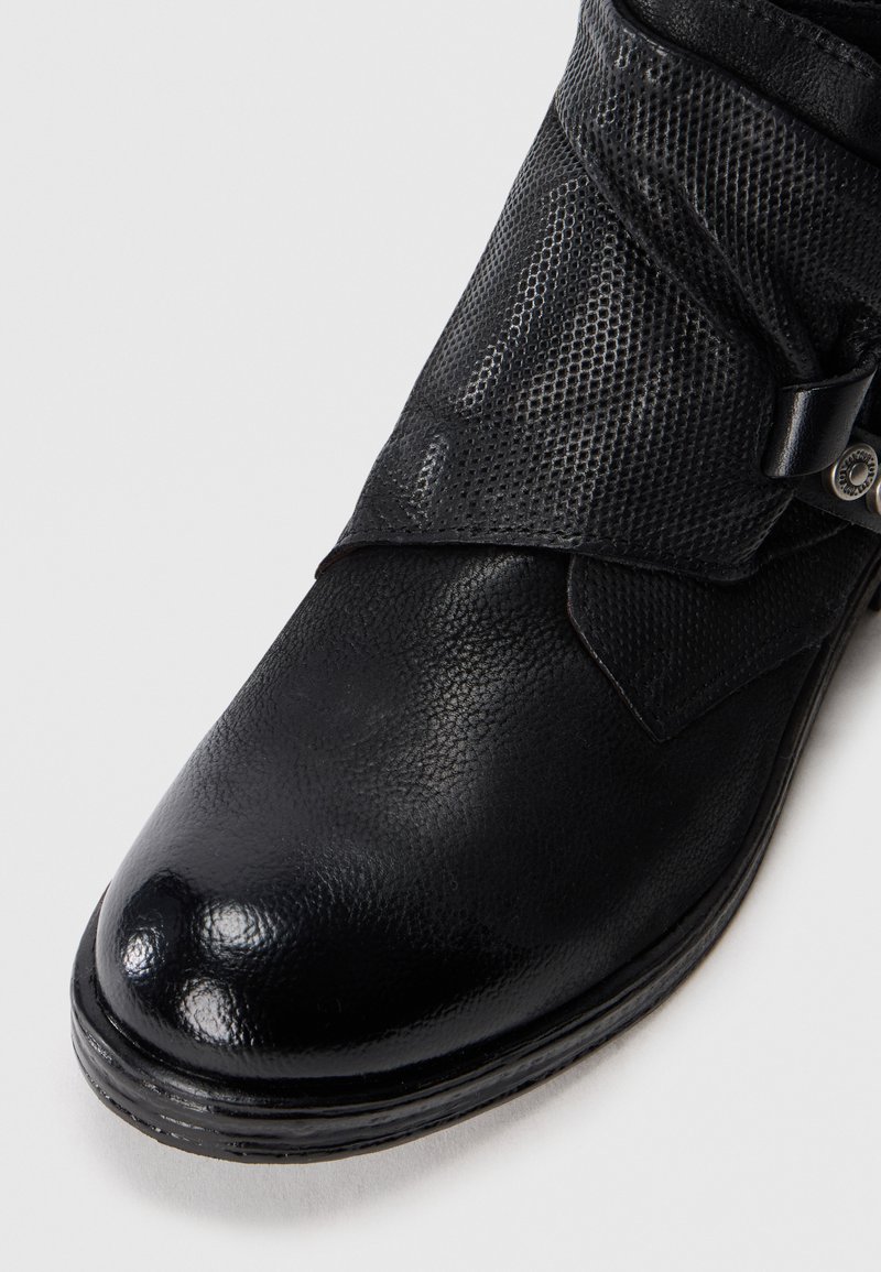 Bota de cuero negro con detalle de hebilla en la correa texturada sobre un fondo claro, mostrando la parte superior frontal y lateral.