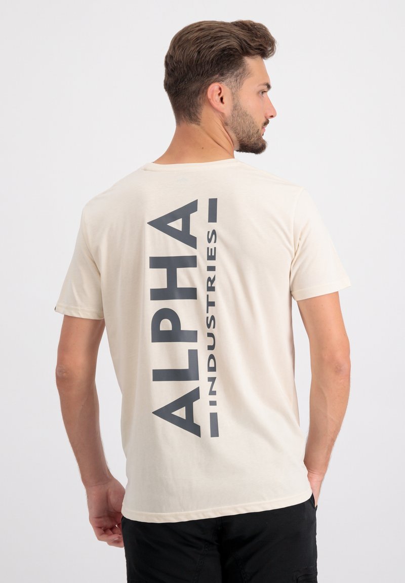 Beige T-Shirt mit kurzen Ärmeln, das auf der Rückseite ein markantes dunkelgraues "ALPHA INDUSTRIES"-Grafikdesign aufweist, mit rundem Ausschnitt und weicher Textur.