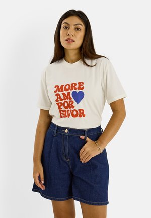 Femme aux longs cheveux foncés portant un T-shirt blanc avec un texte rouge et un cœur bleu, assorti à un short en denim foncé taille haute.