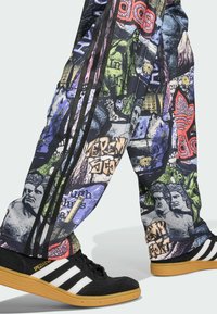 Mehrfarbige grafische Sweatpants mit Collage-Mustern; verfügen über markante Bilder und schwarze Seitenstreifen; kombiniert mit schwarz-weißen Turnschuhen.