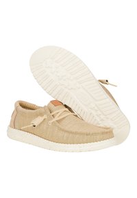 Zapatos de lona beige con un acabado texturizado, cordones neutros y una suela ligera moteada. Incluye un acento de cuero marrón cerca del tobillo.