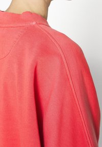 Sudadera de color coral con cuello acanalado y textura suave, con costuras visibles y un corte relajado. La vista posterior muestra las costuras de los hombros.