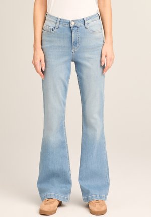 Woman wearing light blue high-waisted flare jeans and beige shoes, standing with hands relaxed by sides.