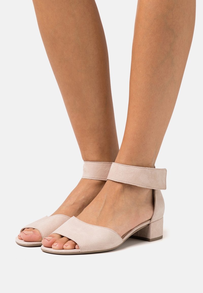 Pieds portant des sandales ouvertes en daim rose clair avec des brides à la cheville et des talons carrés bas sur un fond blanc uni.