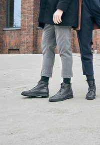 Stivaletti in pelle nera con lacci, suole testurizzate e dettagli in argento. Indossati con pantaloni grigi e blu navy su misura, che mostrano un design moderno.