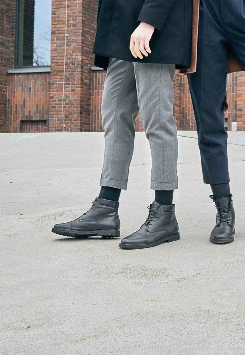 Stivaletti in pelle nera con lacci, suole testurizzate e dettagli in argento. Indossati con pantaloni grigi e blu navy su misura, che mostrano un design moderno.