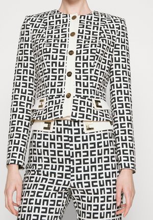 Chaqueta y pantalones con patrón en blanco y negro, botones y hebillas de cinturón dorados, usados por una persona de pie frente a un fondo liso.