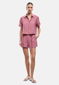Camicetta corta a maniche corte e pantaloni abbinati in un tessuto leggero color rosa chiaro, con bottoni dorati e una cintura in vita. Sandali dal design minimalista.