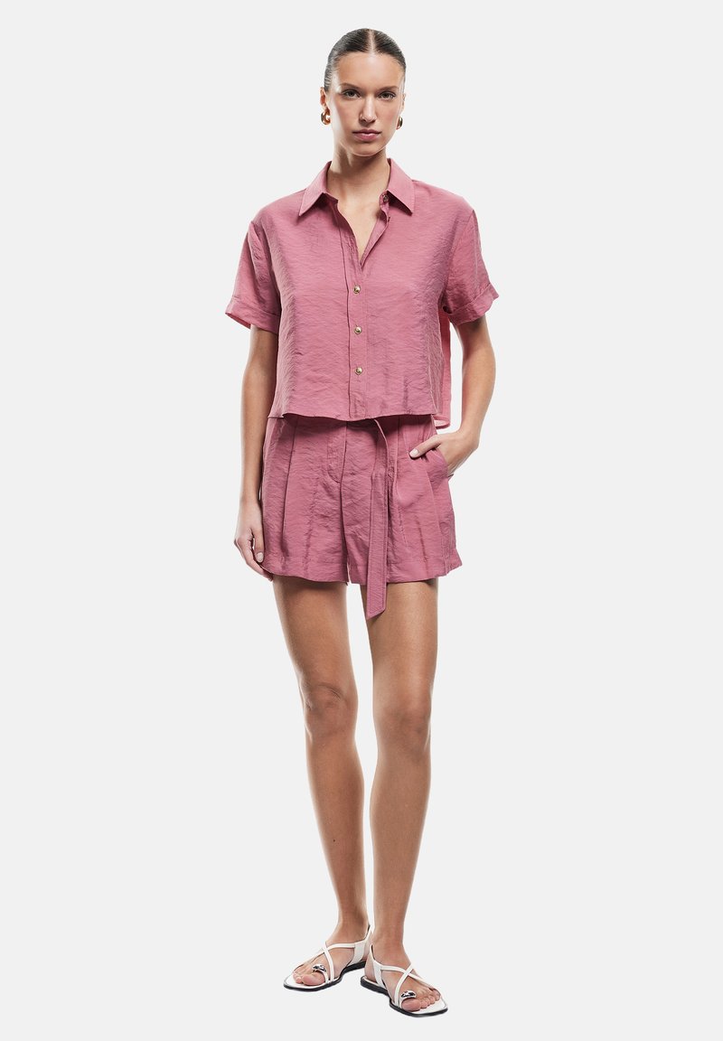 Camicetta corta a maniche corte e pantaloni abbinati in un tessuto leggero color rosa chiaro, con bottoni dorati e una cintura in vita. Sandali dal design minimalista.