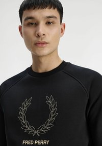 Felpa nera con collo a coste, decorata con una corona di alloro ricamata in oro e il logo "FRED PERRY" sul davanti. Texture liscia.