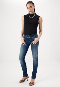 Top negro sin mangas, jeans ajustados azules con desgaste, cierre de dos botones y zapatos de tacón alto marrones con acentos de tachuelas.