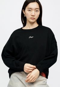 Femme aux longs cheveux noirs portant des boucles d'oreilles en or, un sweat-shirt noir avec le texte "Boss" et un pantalon clair, posant devant un fond blanc.
