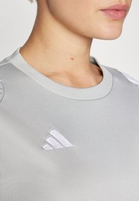 Lys grå treningsskjorte med korte ermer, rund hals, med en hvit brodert logo på brystet og en glatt, myk tekstur.