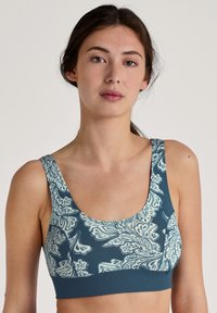 Teal sportbh met een wit paisleypatroon, brede banden en een gladde textuur. Het ontwerp bedekt de voorkant en de zijkanten.