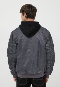 Szara kurtka bomberka o teksturowanym wykończeniu, z czarnym kapturem, elastycznymi mankietami i dołem. Zawiera subtelne detale przeszyć na rękawach.